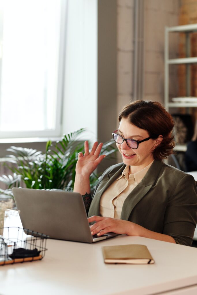 Woman Wearing Blazer and Glasses Waving at Screen While Using MacBook