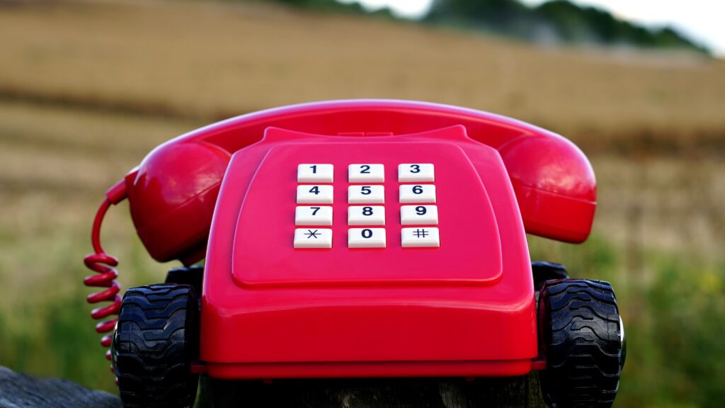 Red Rotary Phone With Hashtag Pound Sign on Keypad with Black Wheels Near Brown Grasses during Day Time
